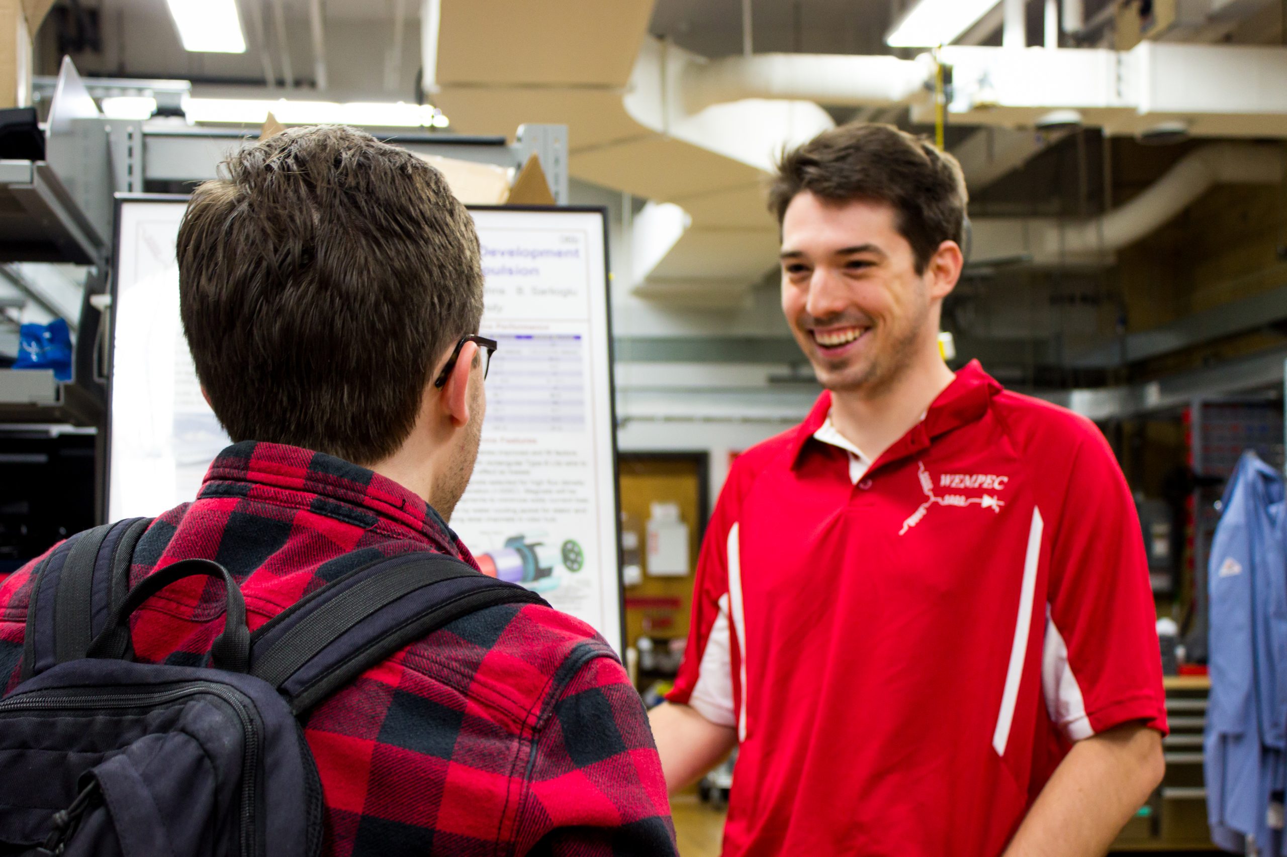 A student in a red WEMPEC polo talking to a business partner about a poster.